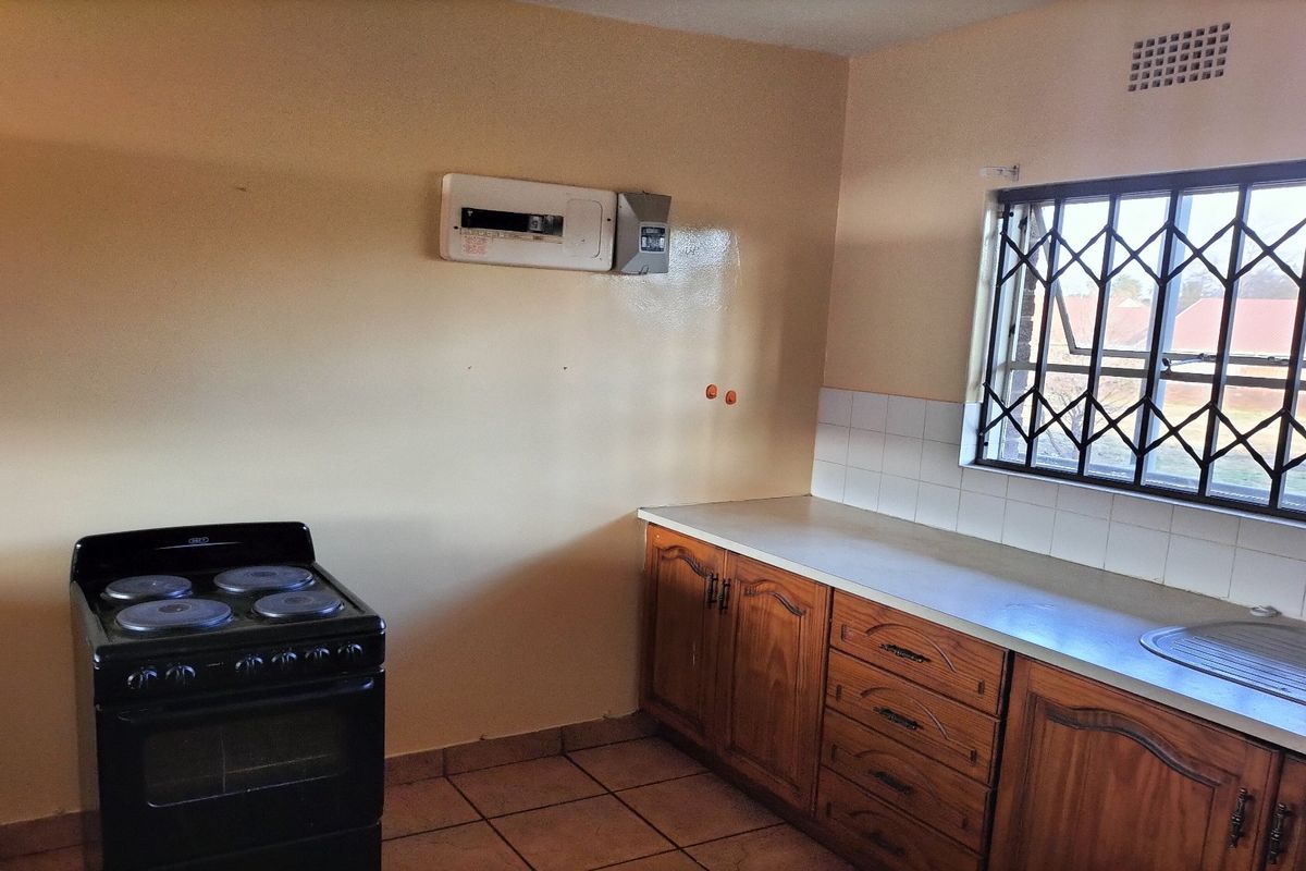 A functional kitchen featuring wooden cabinetry, a tiled backsplash, a stainless steel sink, and a freestanding stove, with a window fitted with security bars.