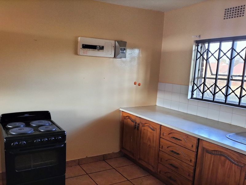 A functional kitchen featuring wooden cabinetry, a tiled backsplash, a stainless steel sink, and a freestanding stove, with a window fitted with security bars.