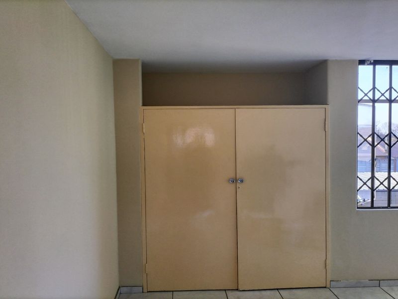 A clean, empty bedroom featuring a built-in cupboard, tiled flooring, and a window fitted with a security gate.