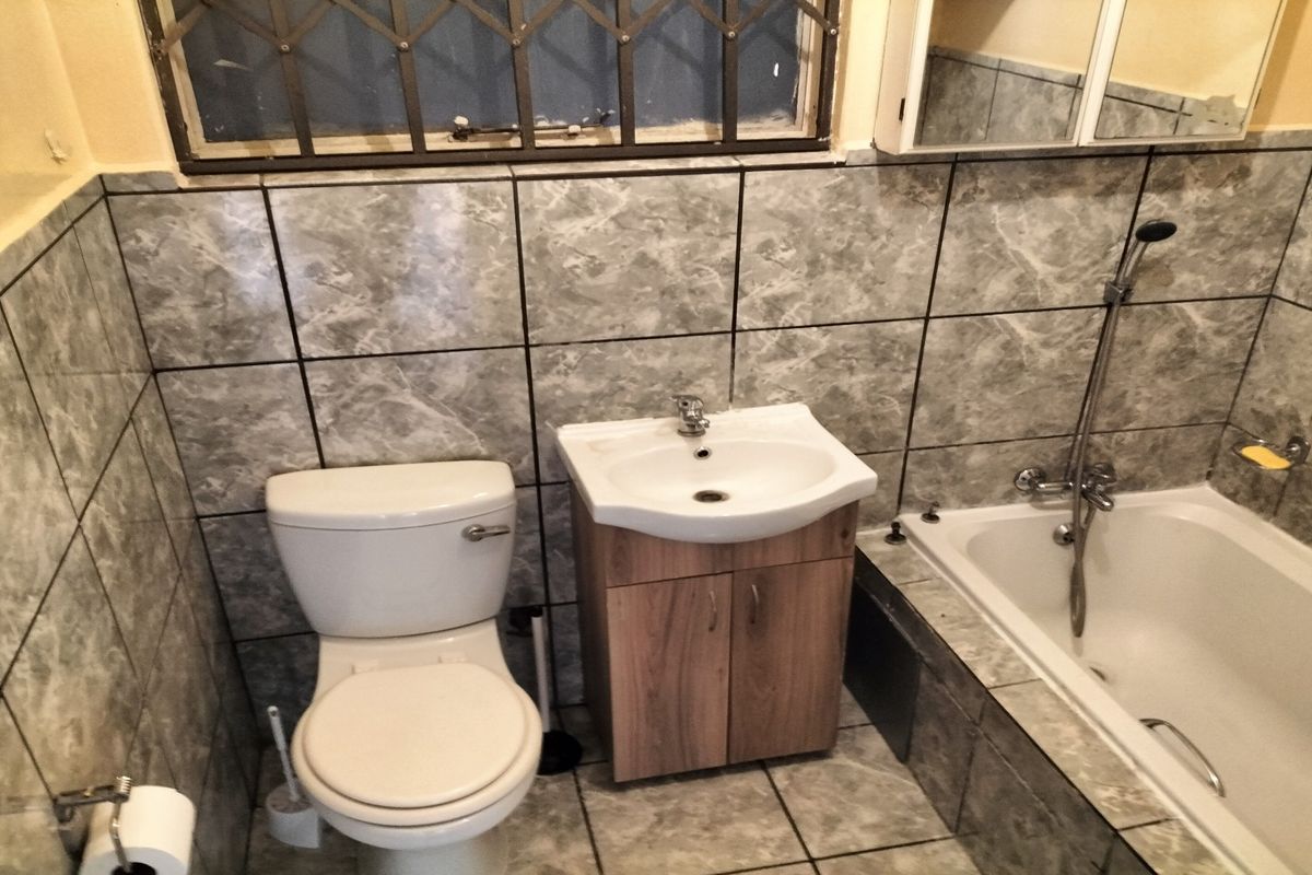 A functional bathroom featuring a bathtub with shower mixer, vanity cabinet with washbasin, toilet, and grey marble-effect wall and floor tiling.