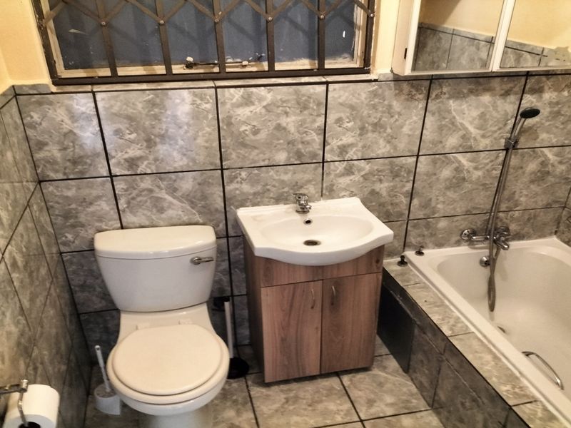 A functional bathroom featuring a bathtub with shower mixer, vanity cabinet with washbasin, toilet, and grey marble-effect wall and floor tiling.