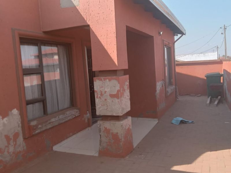 Front exterior view of the house featuring a paved patio area, showing significant wear to the exterior wall paint and plaster finishes.