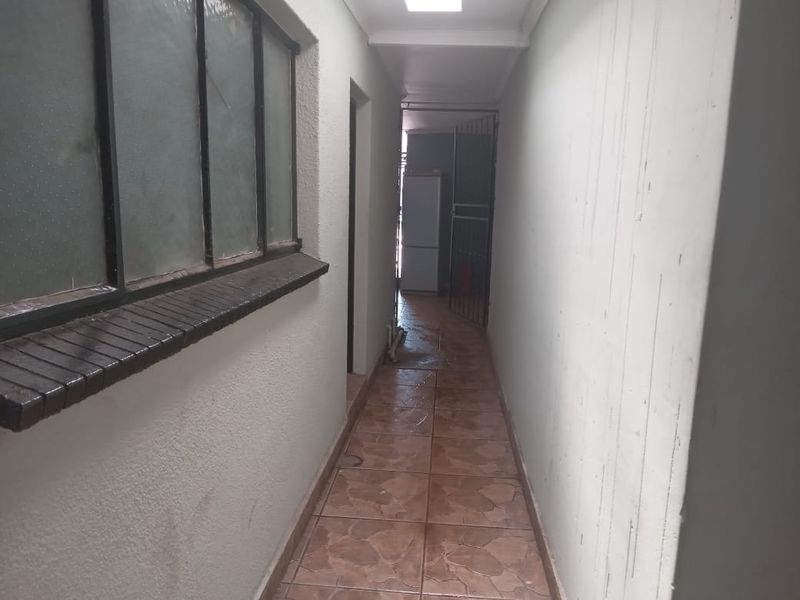 A narrow, tiled hallway with metal-framed windows and recessed lighting, showing signs of wear on the wall surfaces.