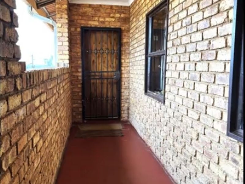 A covered patio area featuring exposed brick walls, a secure entrance door, and a painted concrete floor.