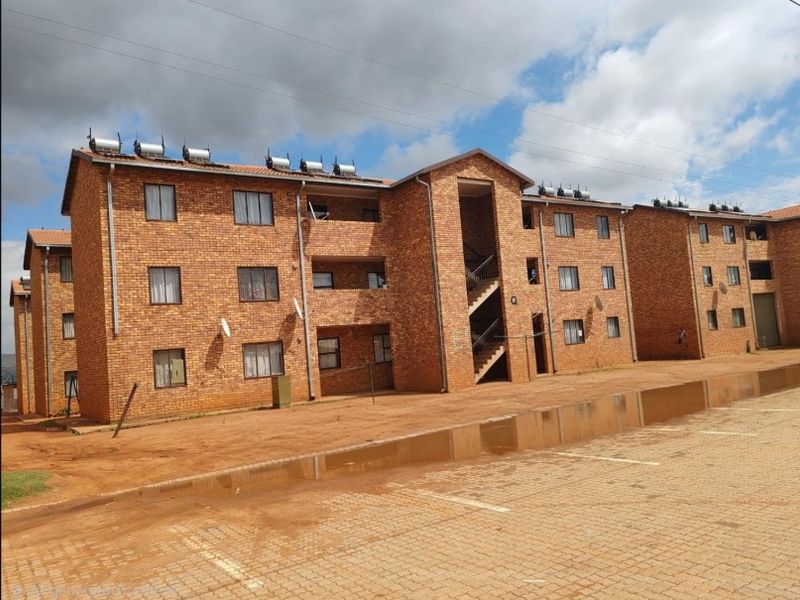 Exterior view of a multi-story face brick apartment complex featuring external staircases, solar water heating systems, and a paved parking area.