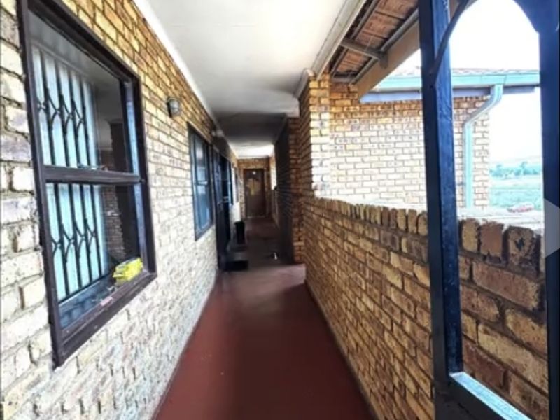 An exterior covered walkway featuring exposed brick walls, security bars on windows, and painted concrete flooring, providing access to the flat units.