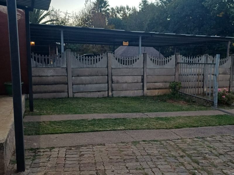 Front exterior view of the cottage featuring a paved driveway, covered carport, and a secure precast concrete boundary wall with a metal gate.