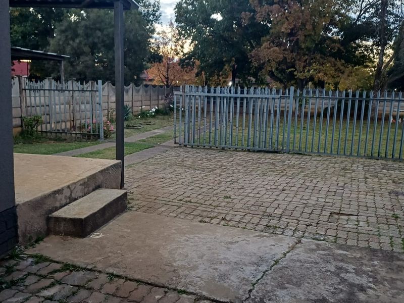 Rear exterior area featuring a paved courtyard, covered patio space, and secure metal fencing, offering potential for outdoor landscaping and improvement.