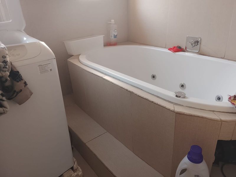 A bathroom featuring a built-in jetted bathtub with tiled surround and wall-mounted fixtures, showing signs of wear on the tile edges.