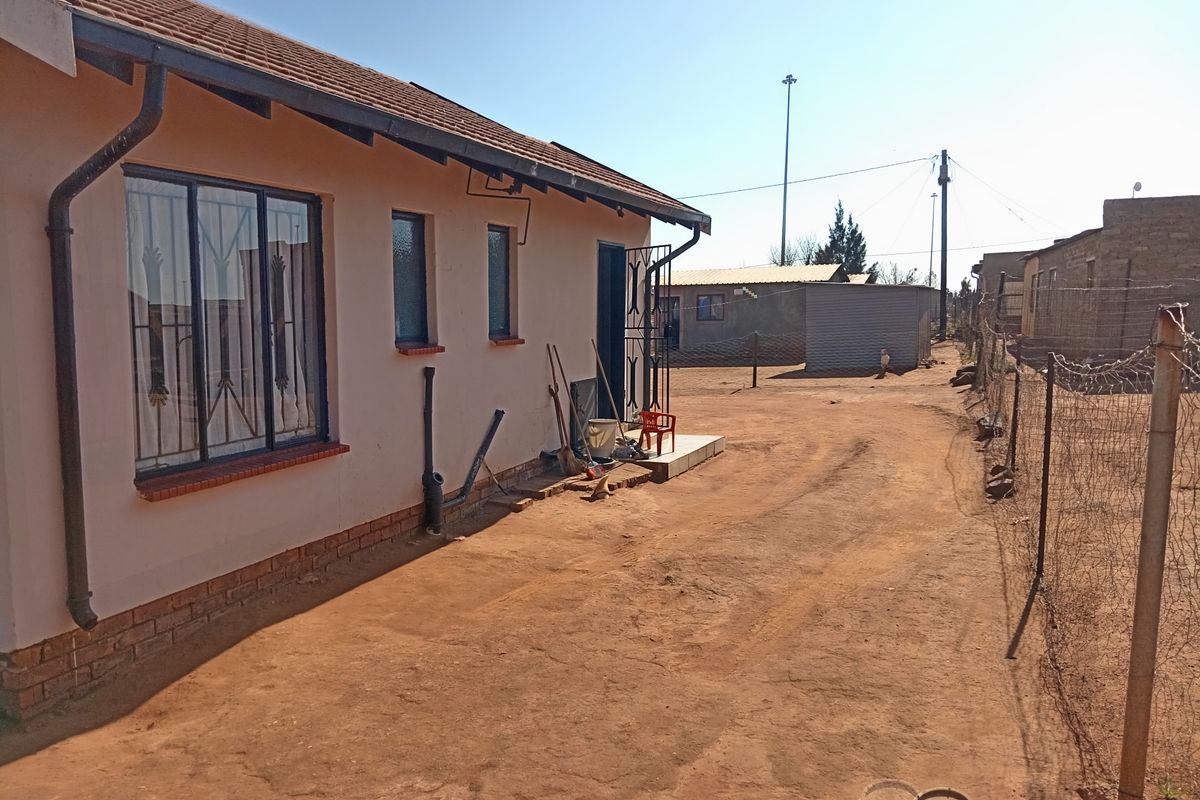 Rear exterior view of the house featuring a tiled roof, security bars on windows, and an unpaved yard area with perimeter wire fencing.