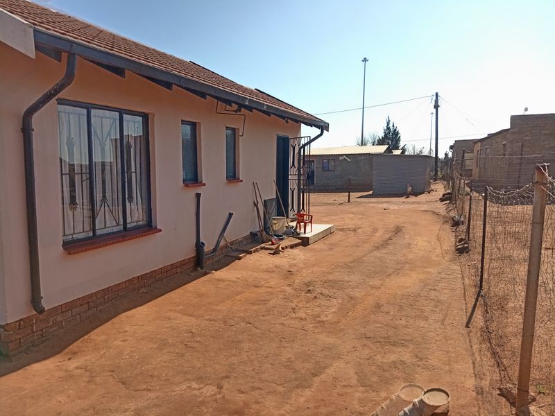 Rear exterior view of the house featuring a tiled roof, security bars on windows, and an unpaved yard area with perimeter wire fencing.
