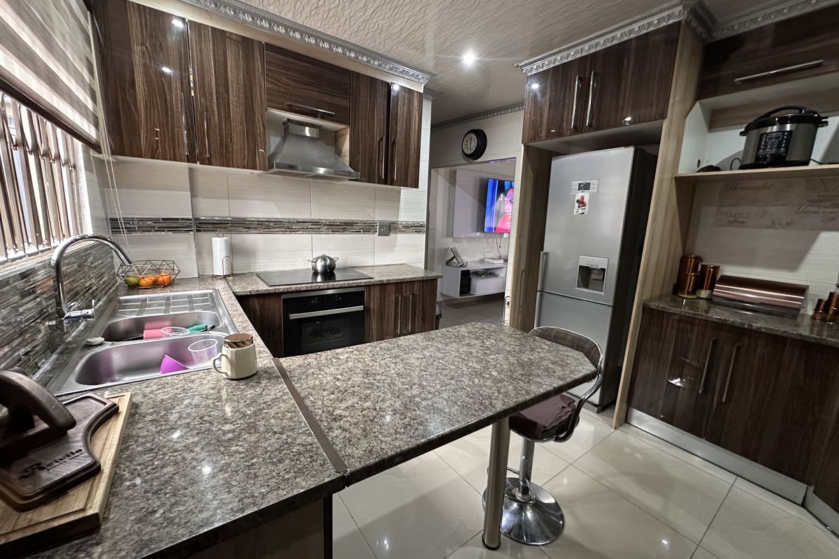 Modern kitchen featuring high-gloss wood-finish cabinetry, granite countertops, integrated appliances, and a breakfast bar, complemented by tiled flooring and a decorative ceiling.
