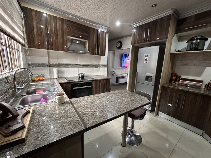 Modern kitchen featuring high-gloss wood-finish cabinetry, granite countertops, integrated appliances, and a breakfast bar, complemented by tiled flooring and a decorative ceiling.