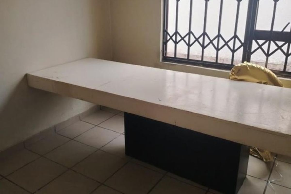 A sparse bedroom featuring a built-in desk, tiled flooring, and a window fitted with a security gate.