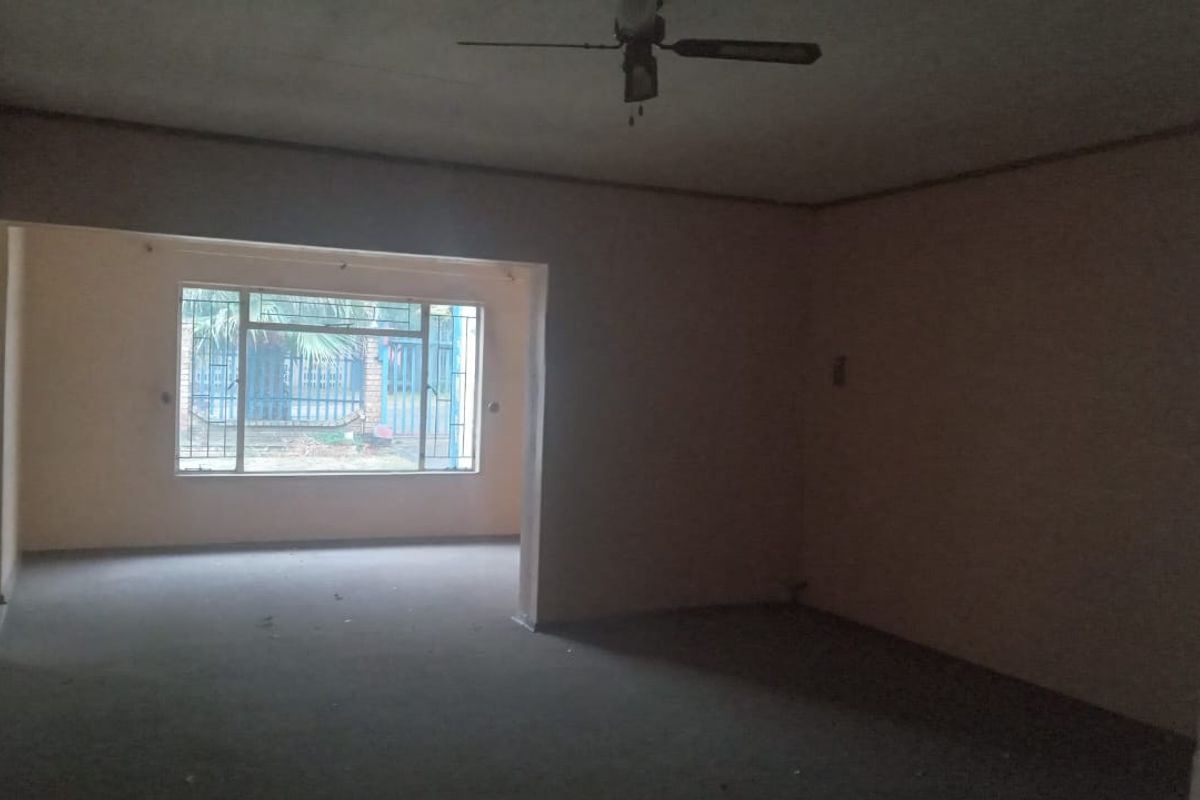 An empty, open-plan living area featuring a ceiling fan and large windows with security bars, offering a blank canvas for renovation.