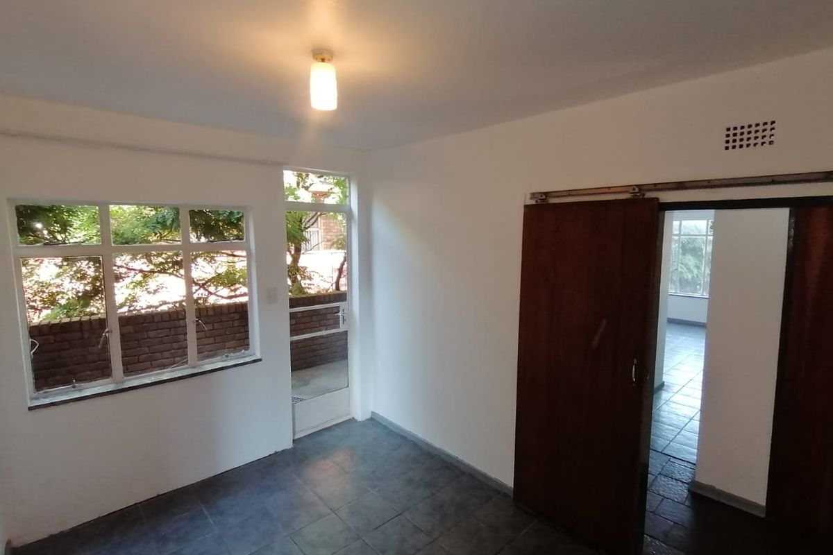 An empty living area featuring tiled flooring, a sliding wooden door, and large windows providing natural light and garden views.