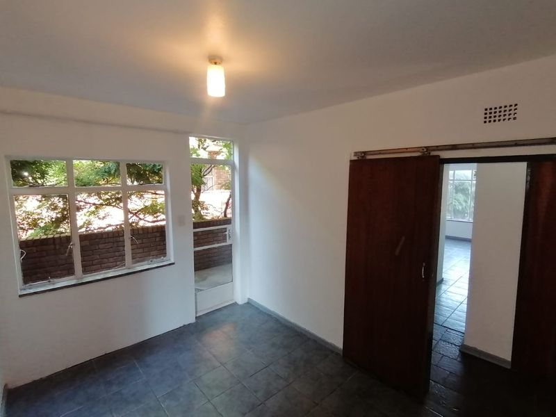 An empty living area featuring tiled flooring, a sliding wooden door, and large windows providing natural light and garden views.