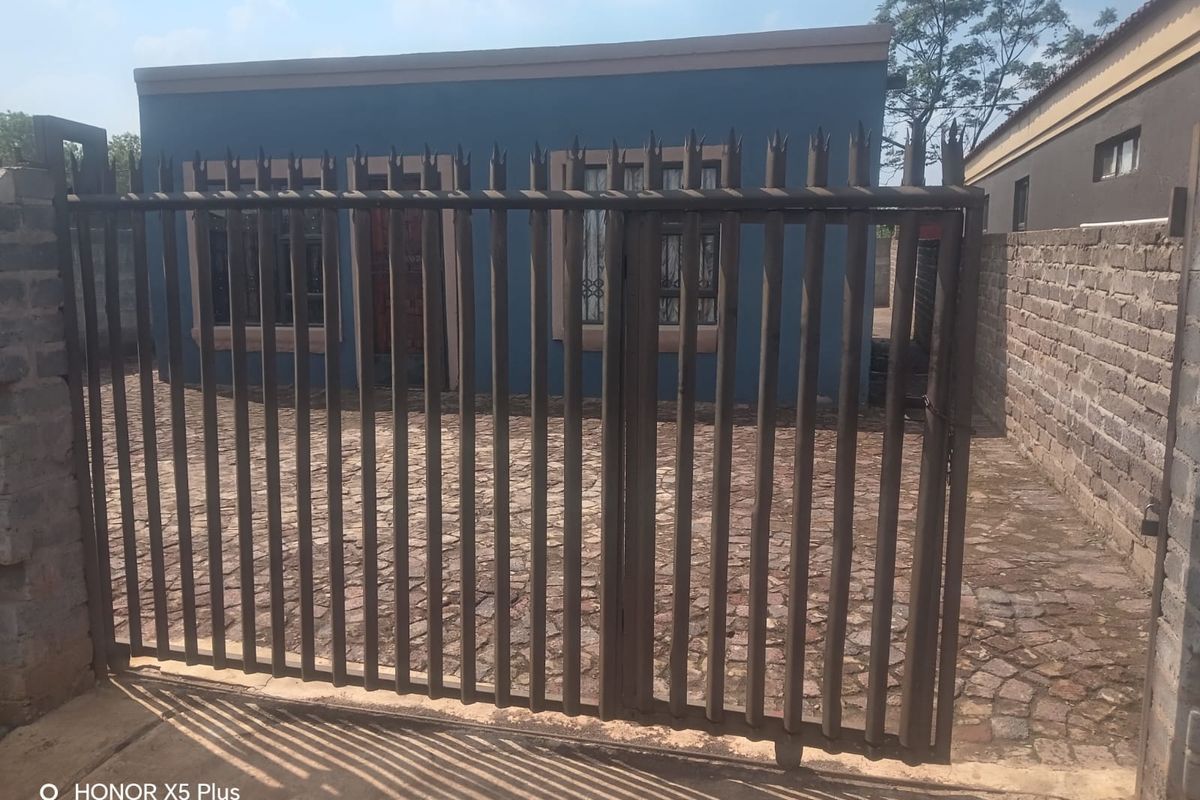Front view of a residential property featuring a metal security gate, paved driveway, and brick boundary wall.