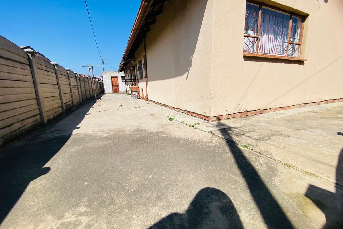 A view of the rear exterior area featuring a concrete paved pathway, precast boundary wall, and the side elevation of the house with security bars.