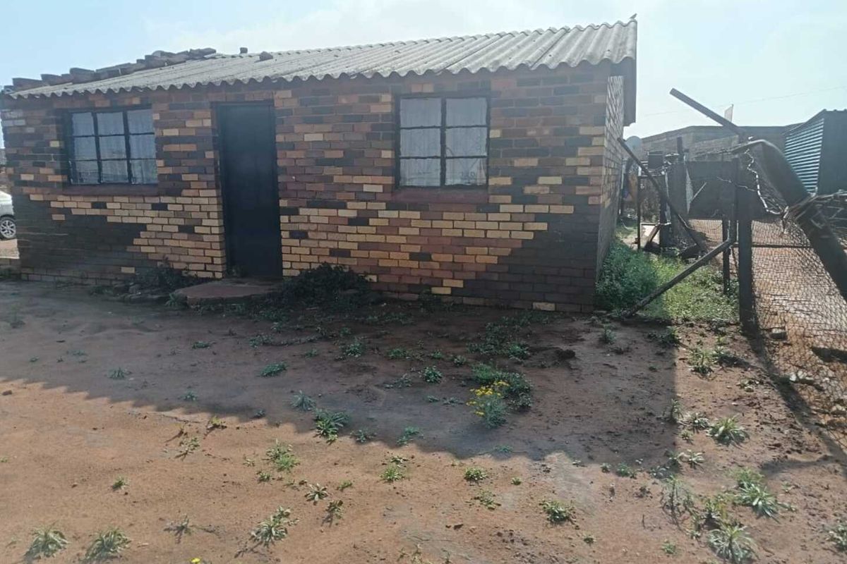 A single-story face brick house featuring a corrugated roof and steel-framed windows, situated on an unpaved plot with perimeter wire fencing.