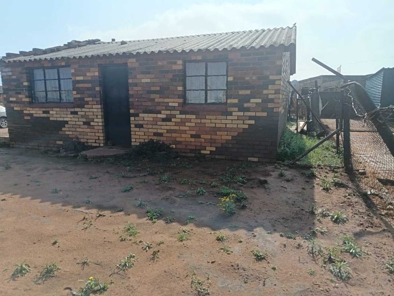 A single-story face brick house featuring a corrugated roof and steel-framed windows, situated on an unpaved plot with perimeter wire fencing.