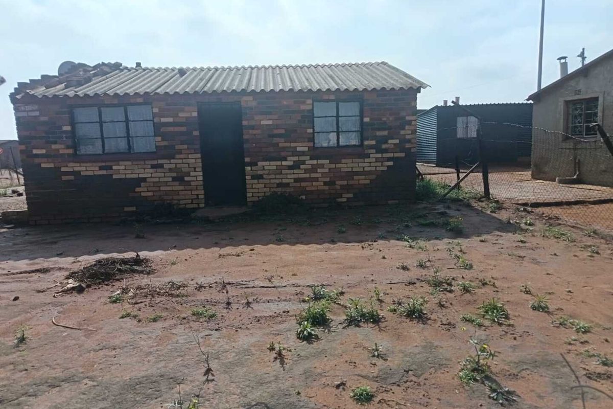 A single-story face brick house featuring a corrugated roof and steel-framed windows, situated on an unpaved plot with perimeter wire fencing.