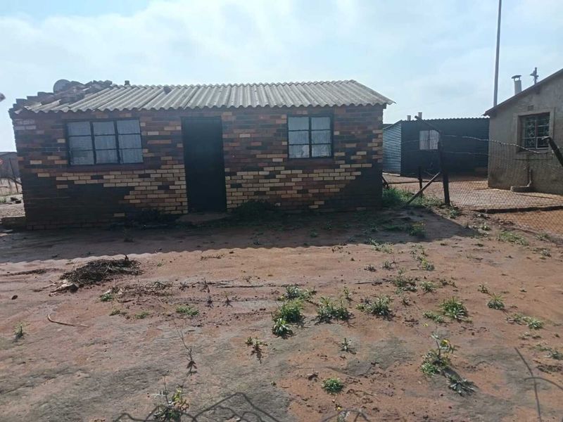 A single-story face brick house featuring a corrugated roof and steel-framed windows, situated on an unpaved plot with perimeter wire fencing.