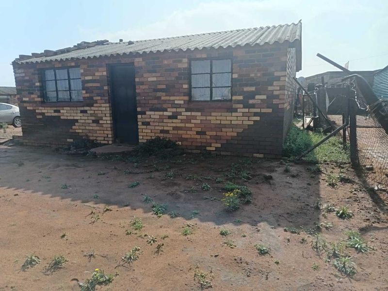 A single-story face brick house featuring a corrugated roof and steel-framed windows, situated on an unpaved plot with perimeter fencing.