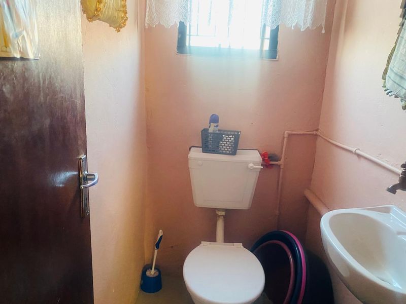 A compact bathroom featuring a standard toilet, washbasin, and window with security bars, showing signs of wear and dated finishes.