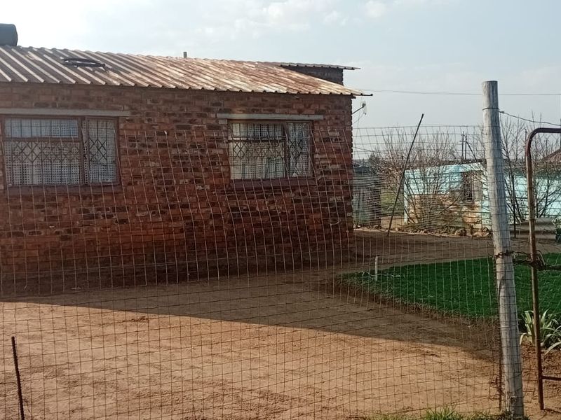 A modest face brick house featuring a corrugated metal roof with a solar water heater, enclosed by wire mesh fencing and a metal gate.