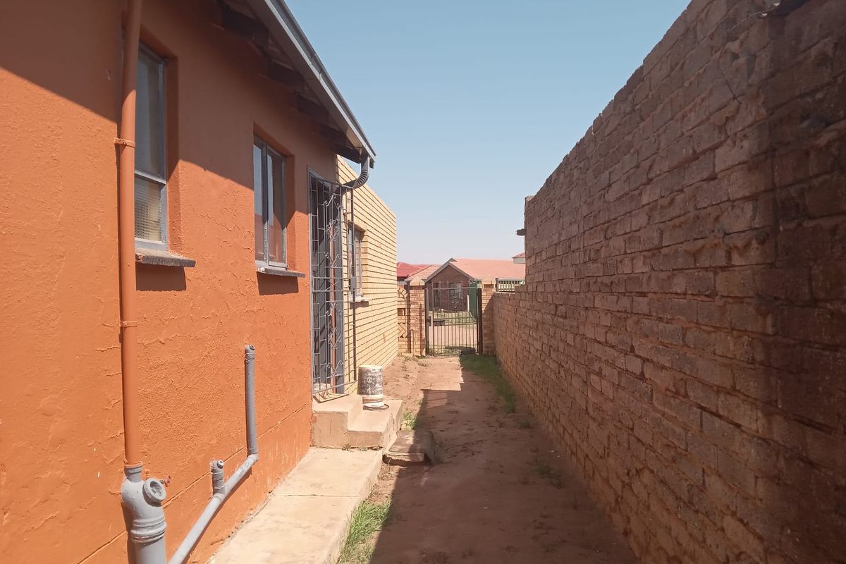 A side exterior view showing a narrow pathway between the house wall and a brick boundary wall with a security gate in the distance.