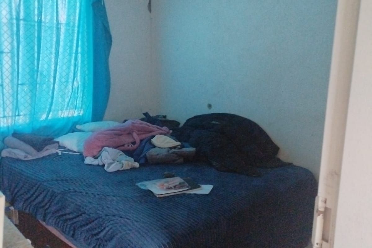 A bedroom featuring tiled flooring and a window with security bars, showing signs of wall plaster damage near the ceiling corner.