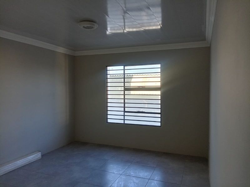 A clean, empty bedroom featuring neutral tiled flooring, a glossy ceiling finish, and a window fitted with security bars.