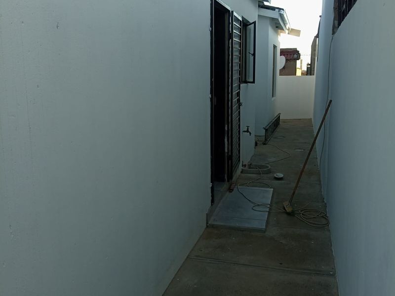 A narrow side passage or rear exterior walkway featuring a concrete path, external water tap, and access to the building via a security gate.