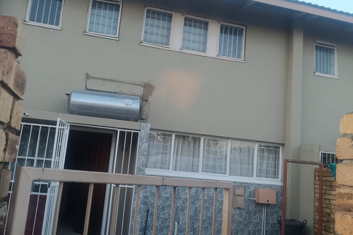 Rear exterior view of the flat featuring a paved courtyard, security gates, and a wall-mounted solar geyser with visible exterior maintenance requirements.