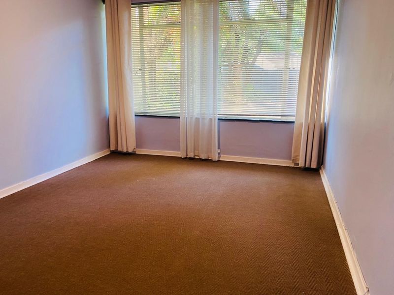 A clean, empty bedroom featuring neutral wall tones, carpeted flooring, and a large window with fitted blinds, offering a blank canvas for personalization.