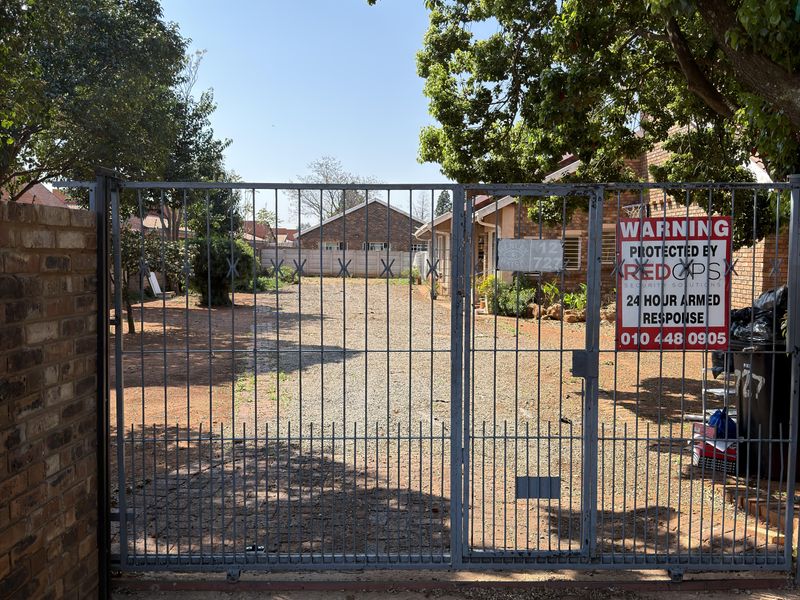 Front entrance view featuring a metal security gate, brick boundary wall, and a gravel driveway leading to the property.