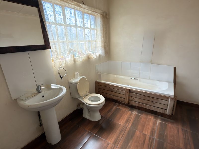 A dated bathroom featuring a pedestal basin, toilet, and bathtub with wood-panel surround, finished with dark tiled flooring and a window with security bars.