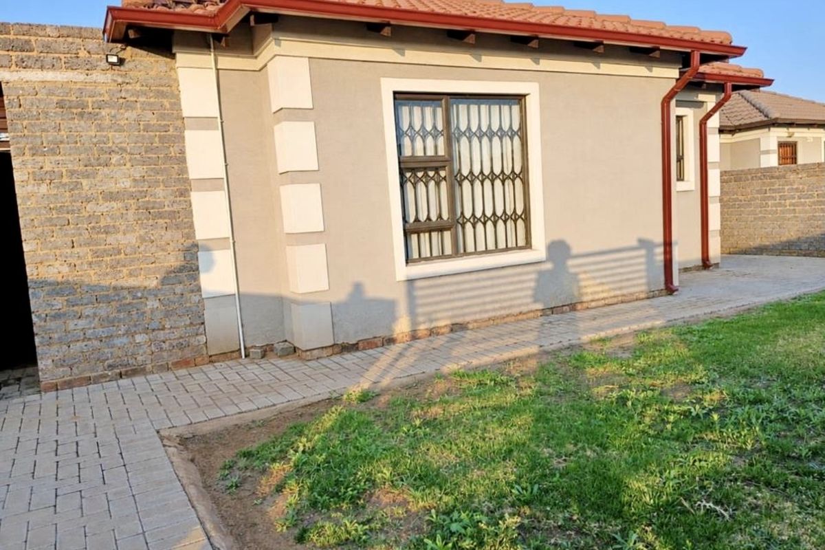 A view of the rear exterior of the house featuring a paved walkway, plastered walls, and a secure window with a metal gate.