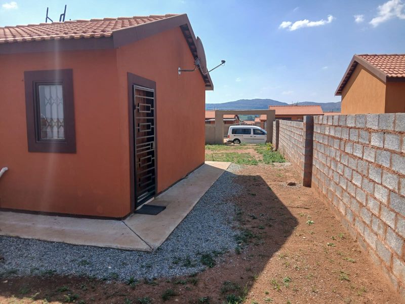 Rear view of the property featuring a terracotta tiled roof, security gate, and a concrete boundary wall with an unfinished garden area.