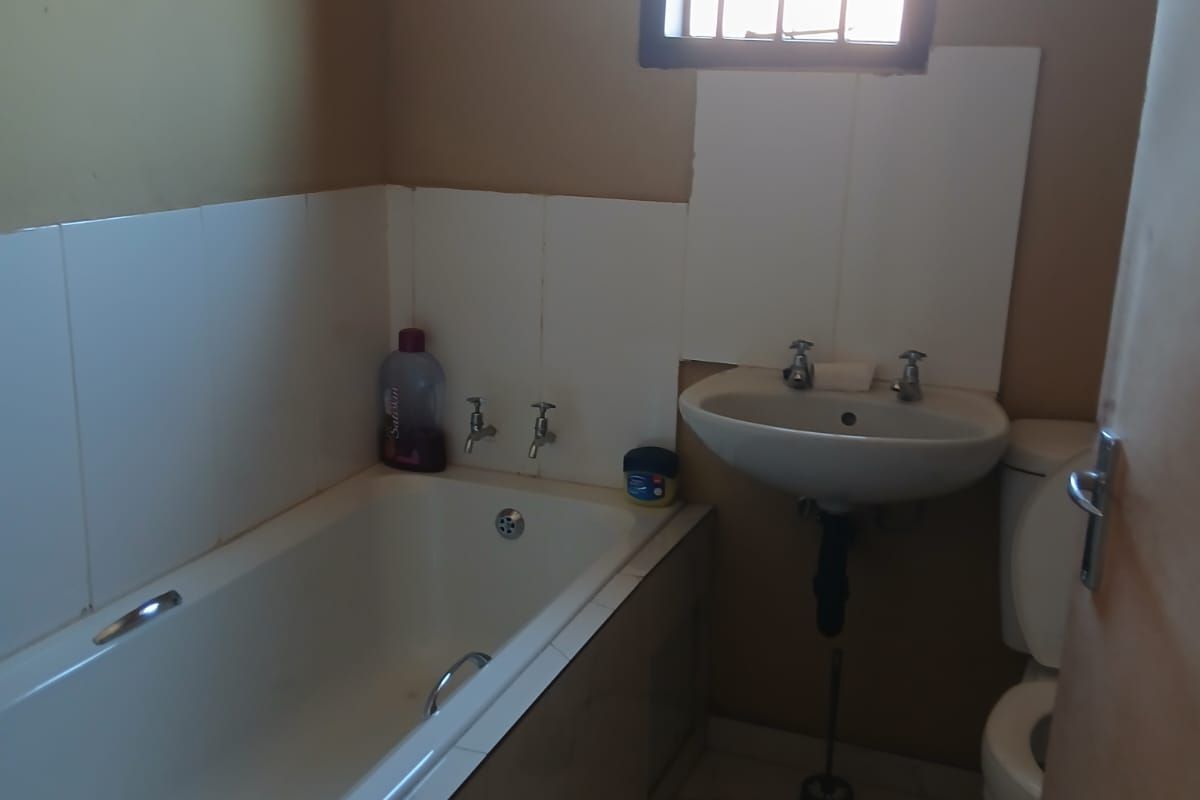 A functional bathroom featuring a standard bathtub, pedestal basin, and toilet, with tiled splashbacks and a window fitted with security bars.