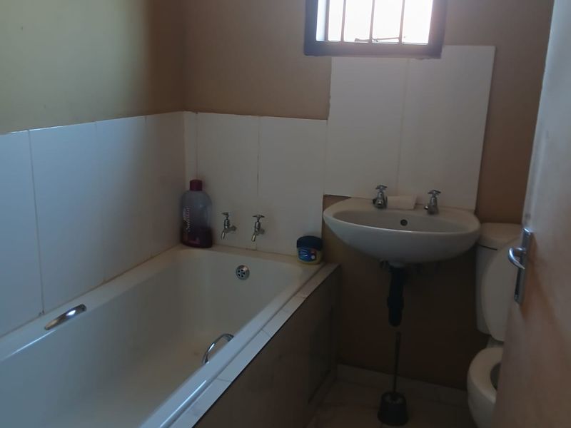 A functional bathroom featuring a standard bathtub, pedestal basin, and toilet, with tiled splashbacks and a window fitted with security bars.