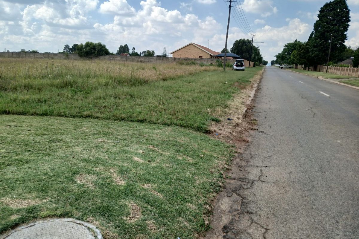 A street-level view of a vacant residential plot featuring grass coverage, a perimeter wall, and adjacent utility infrastructure.
