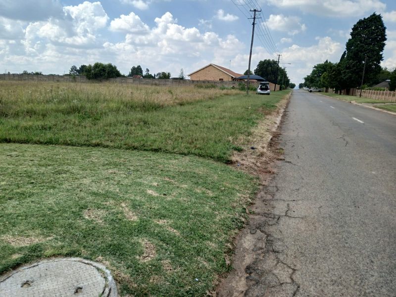 A street-level view of a vacant residential plot featuring grass coverage, a perimeter wall, and adjacent utility infrastructure.