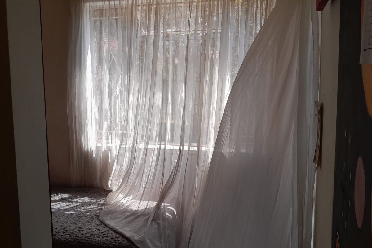 A bedroom interior featuring a window with security bars and sheer curtains, allowing natural light into the space.