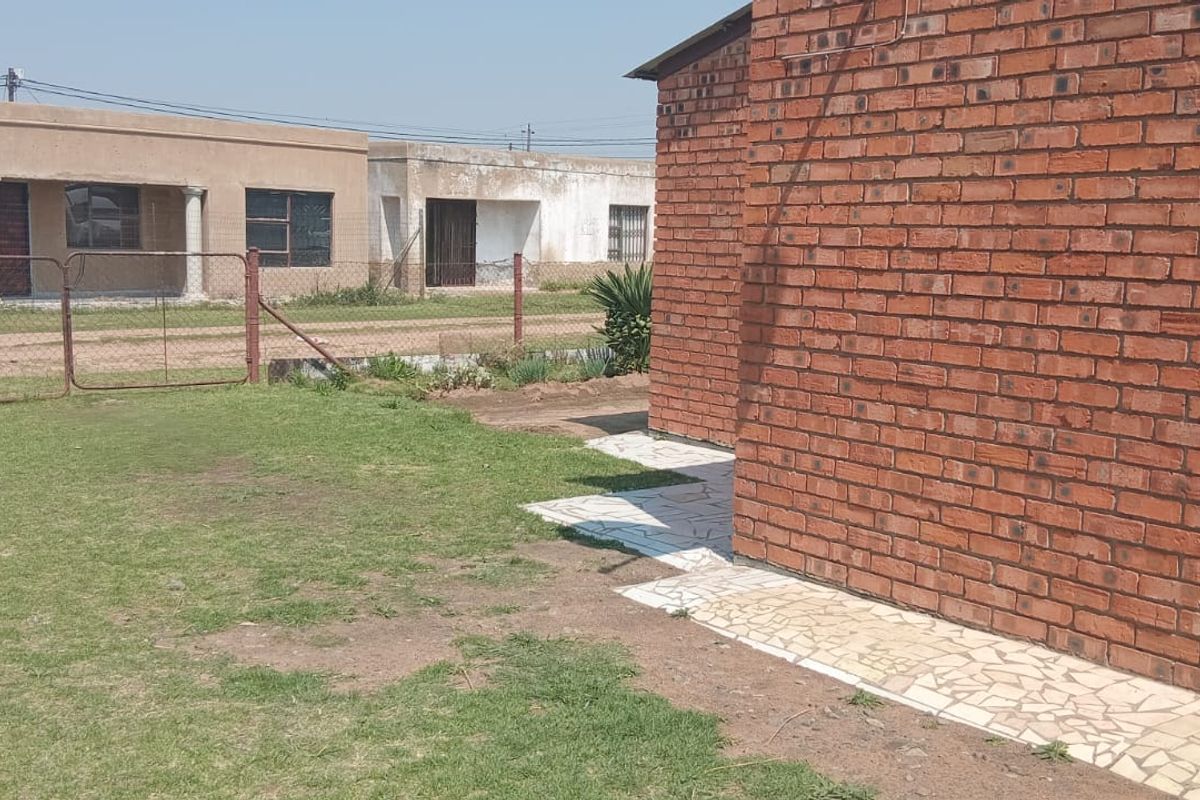 A view of the rear exterior featuring exposed brick wall construction, a crazy paving walkway, and a grassed yard area enclosed by wire fencing.