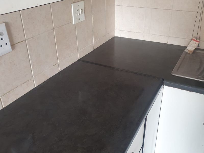 A compact kitchen area featuring white cabinetry, dark laminate countertops, and a tiled backsplash, showing signs of wear and requiring cosmetic updates.