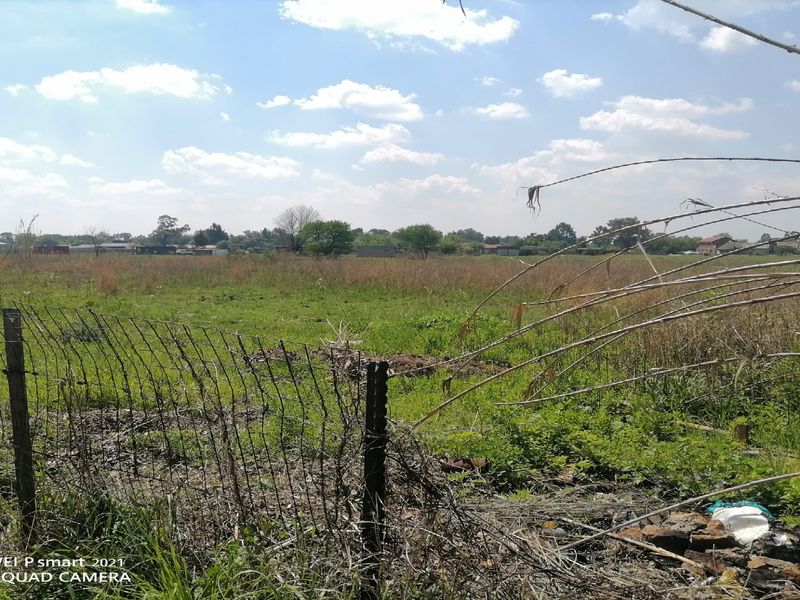 A view of a vacant residential plot featuring overgrown grass and damaged wire mesh perimeter fencing.