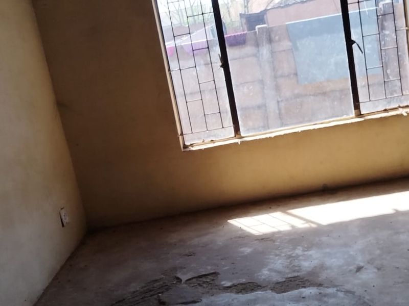 An empty living room featuring concrete flooring, security bars on the windows, and plain wall finishes, requiring cosmetic updates.
