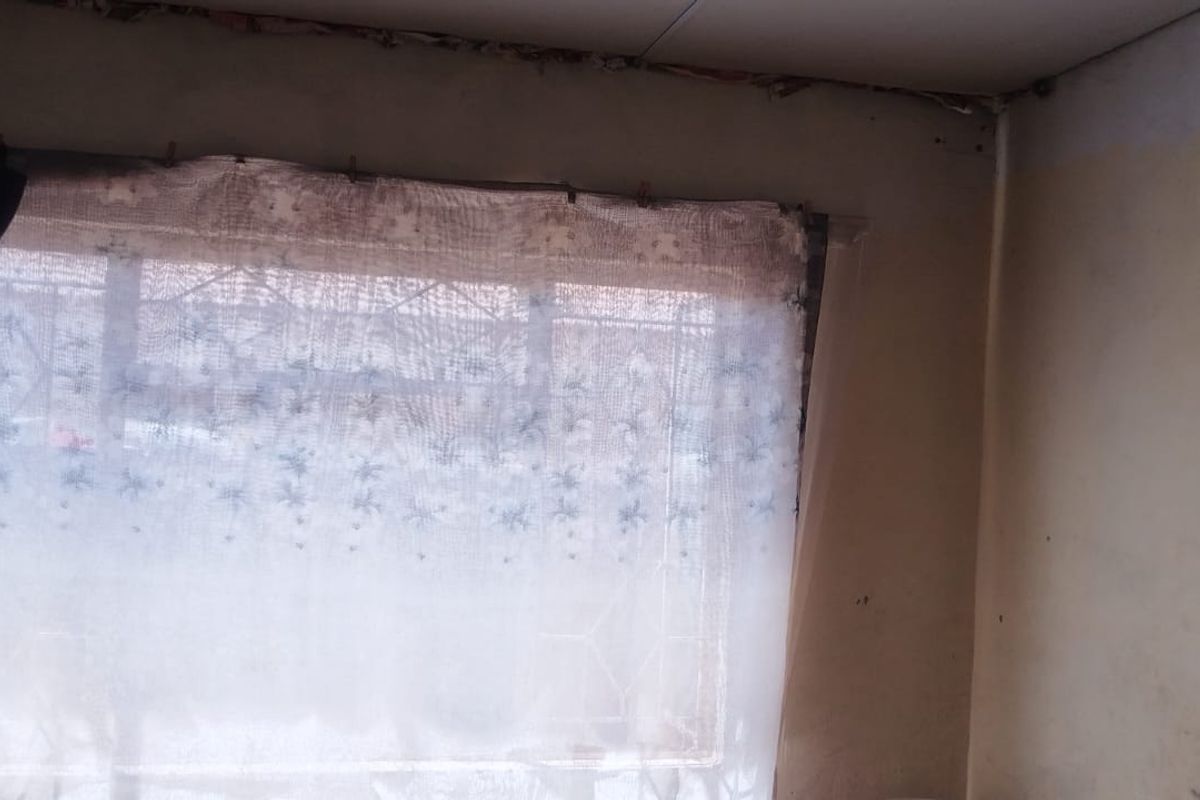 A bedroom interior showing significant wear to the walls and ceiling, featuring a window with security bars.